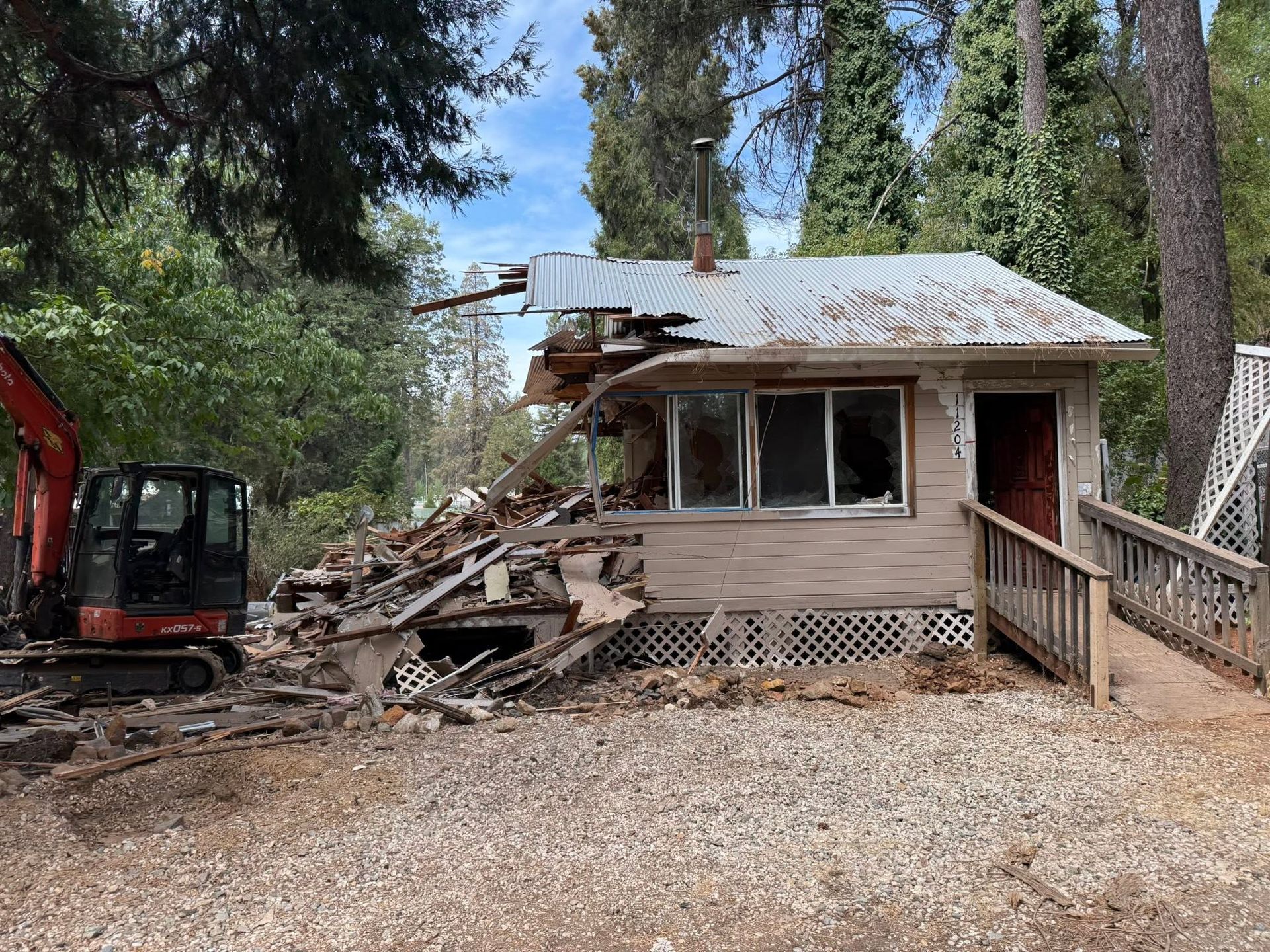 Damaged small house with collapsed roof and debris, beside an excavator in a wooded yard.