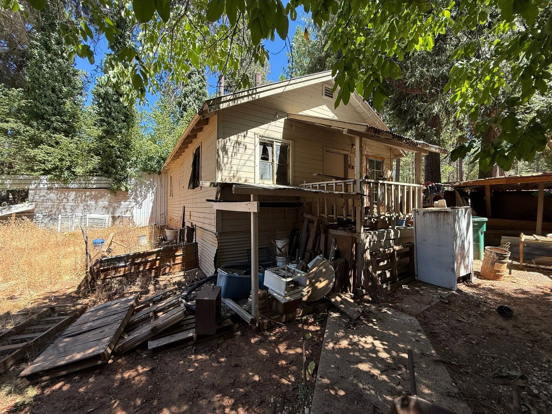 Small house with a porch and cluttered yard under trees, with wood piles and scattered debris nearby