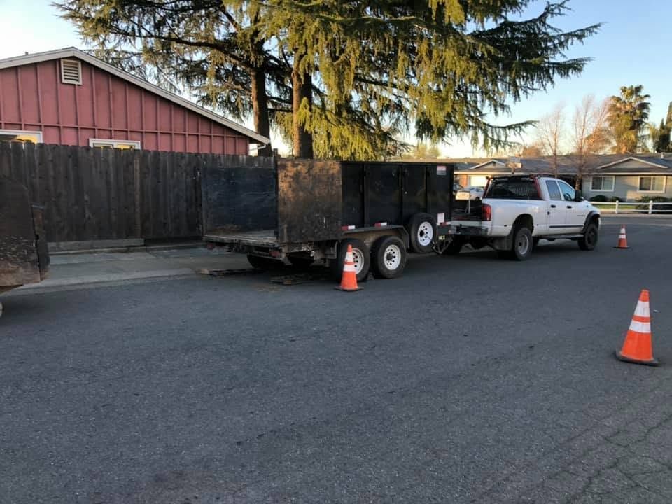 White pickup truck towing a black trailer on a street, with orange cones marking the area.