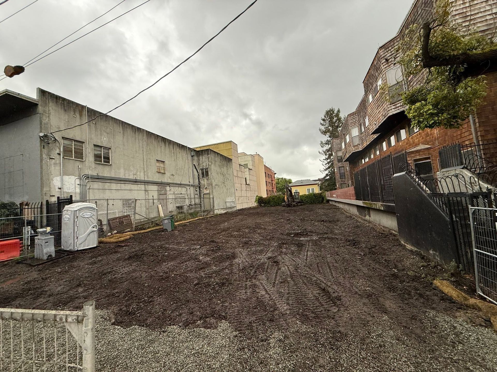 Muddy alley between weathered buildings under cloudy sky, with a mural-covered wall on the right.
