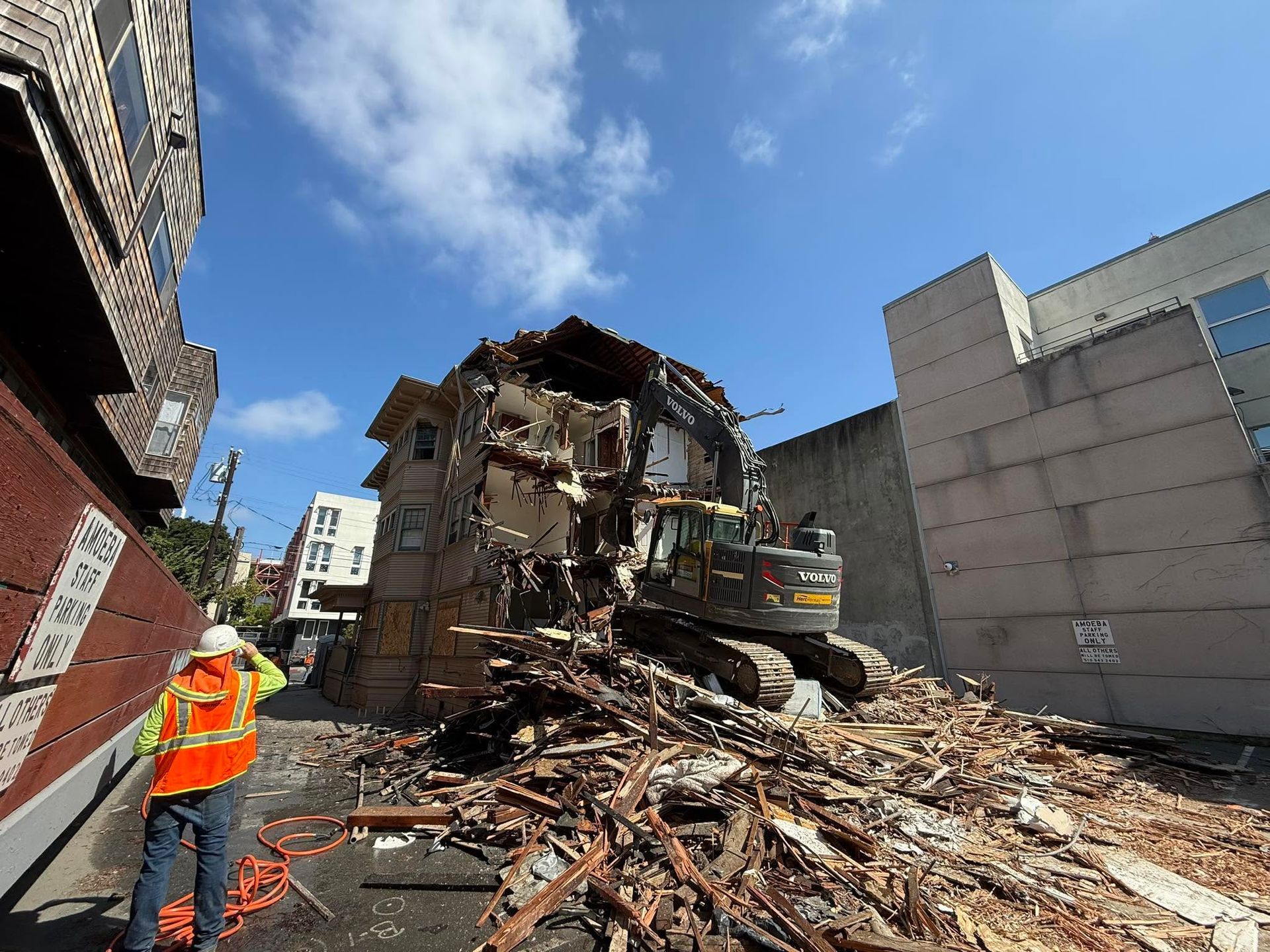 Collapsed building rubble with an excavator and a worker in an orange vest under a blue sky
