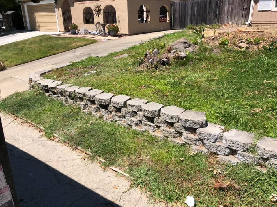 Broken stone border along a grassy slope beside a sidewalk and house driveway.