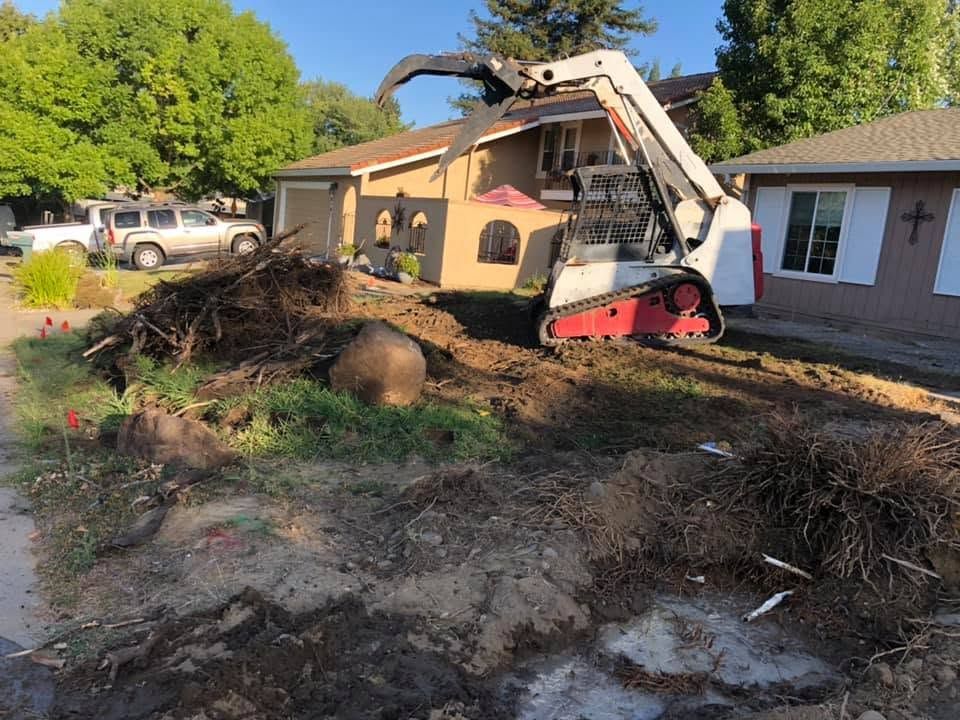 Mini excavator clearing dirt and roots in front of a tan house with trees and a white pickup truck nearby
