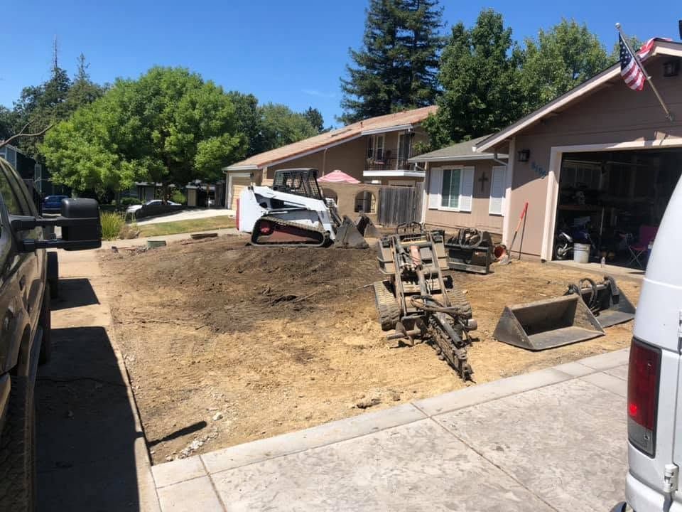 Residential front yard with a skid steer and piles of dirt beside a house and driveway, under bright sunlight