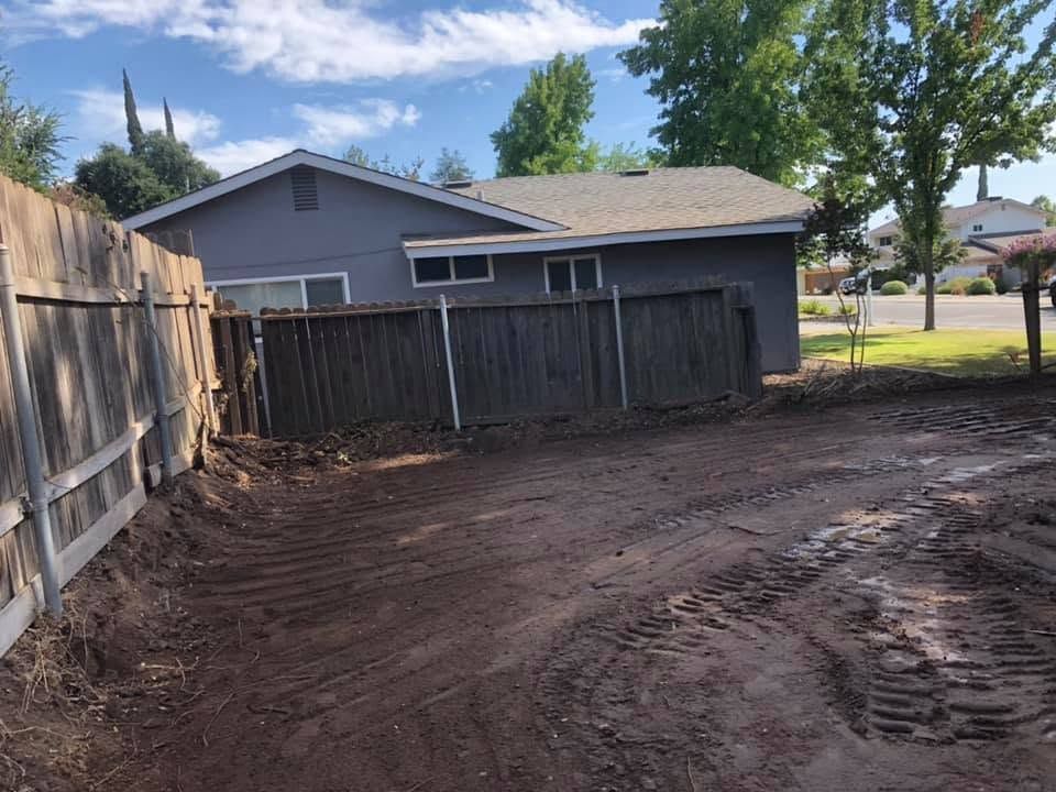 Dirt backyard beside a gray house, with a wooden fence and tire tracks in the soil.