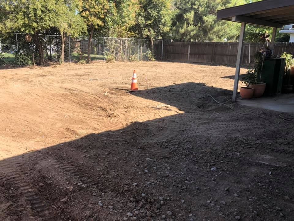 Cleared dirt backyard with a traffic cone and shaded patio beside a wooden fence