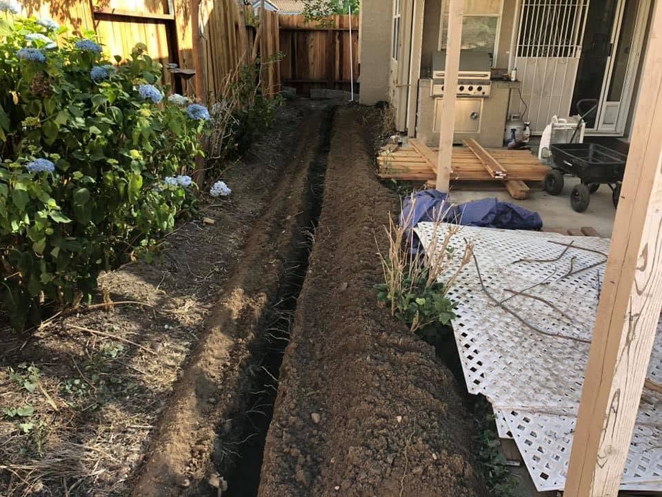 Narrow trench dug along a house and fence, with soil piles beside a patio and garden plants.