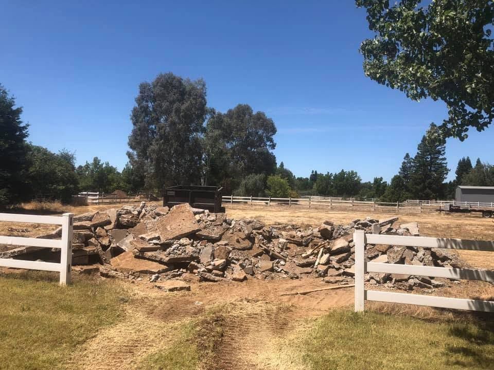 Rural fenced lot with a large pile of rubble and trees under a clear blue sky