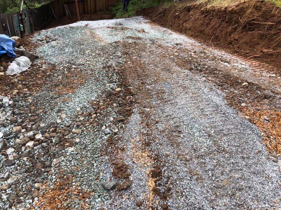 Gravel-covered dirt road with muddy ruts and a small rise, bordered by trees and debris