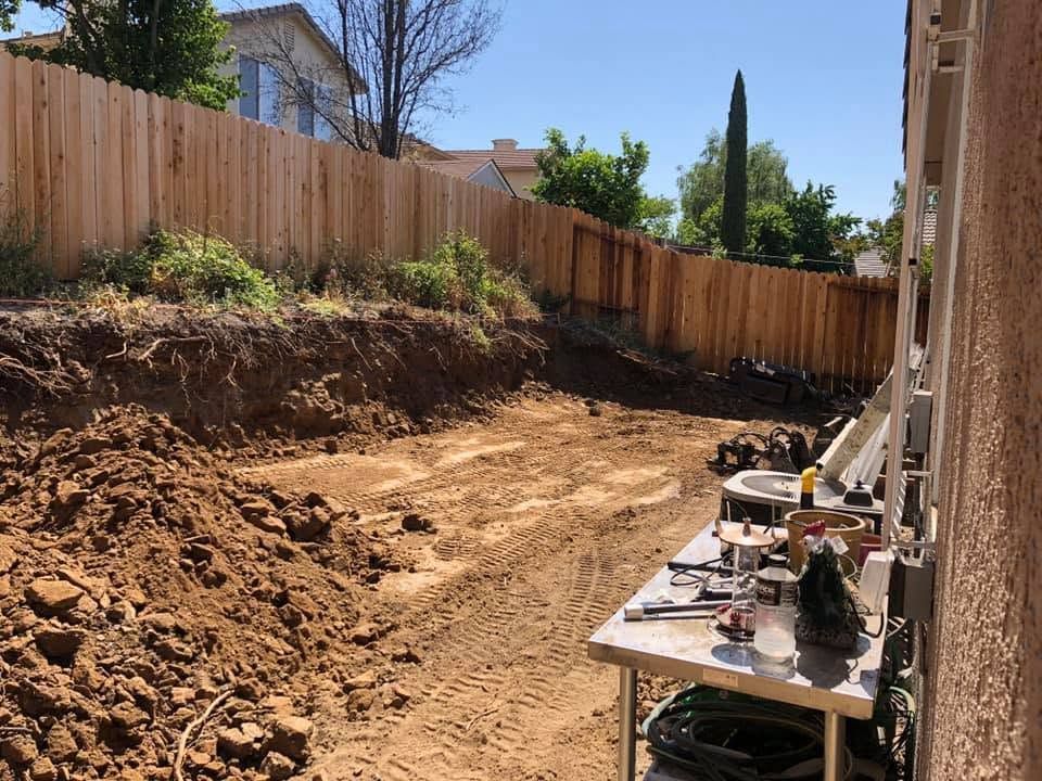 Backyard with excavated dirt slope, wooden fence, and tools on a table beside a house