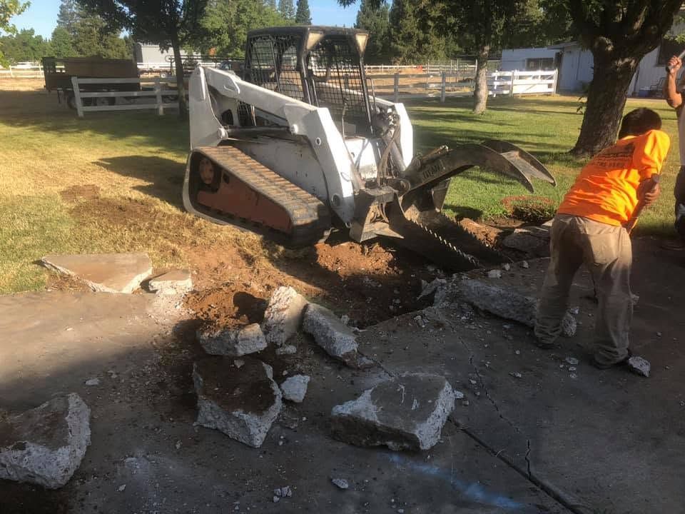 White Bobcat skid steer clearing broken concrete while a worker in an orange shirt stands nearby.