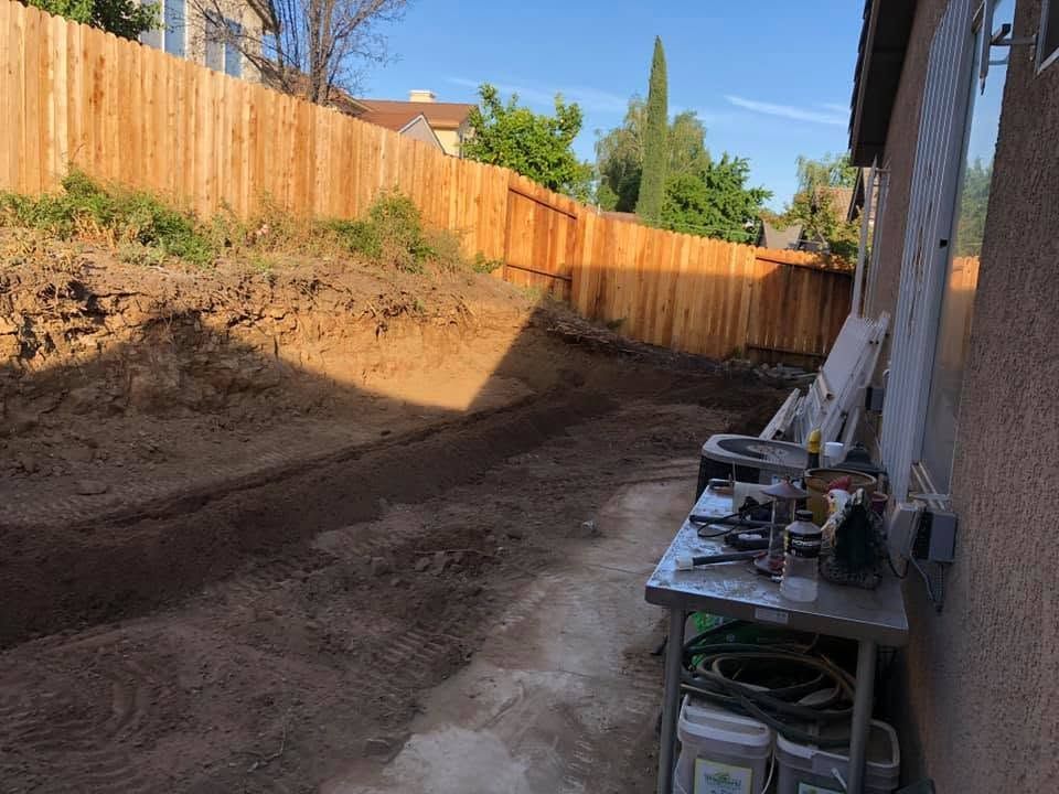 Backyard dirt trench beside house, with wooden fence and a tool table along the wall