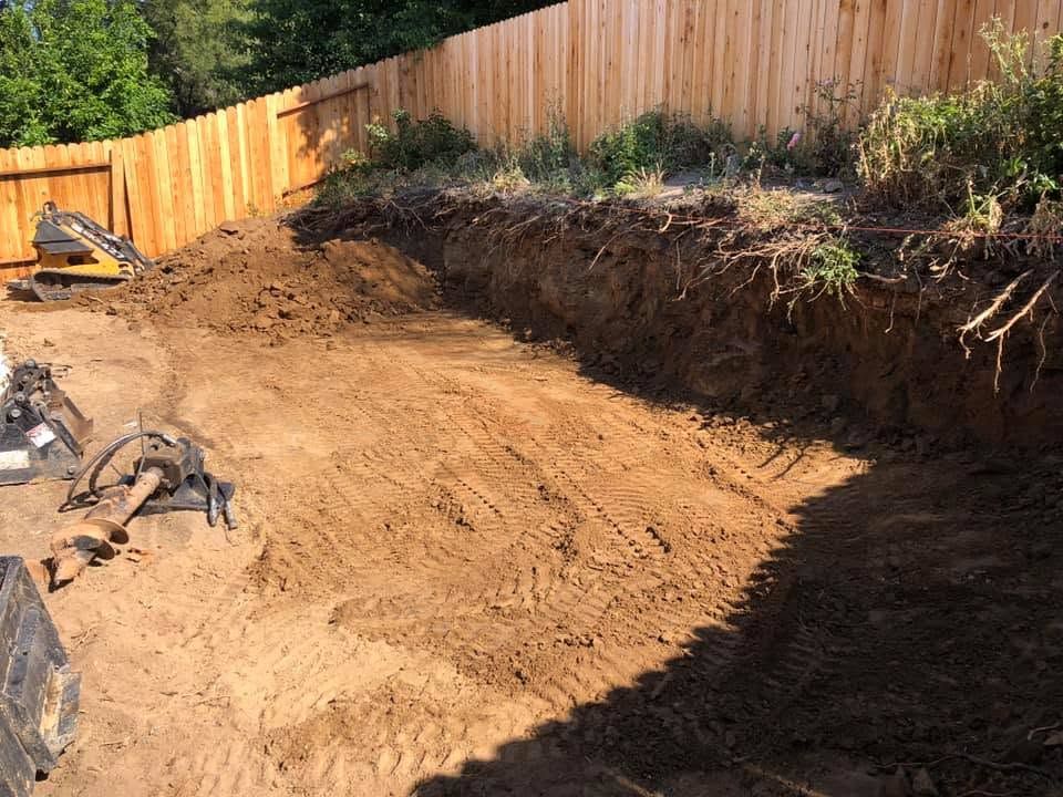 Freshly excavated backyard with a dirt slope, wooden fence, and construction debris in sunlight