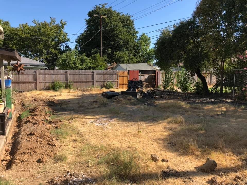 Dry backyard with patchy dirt, a wooden fence, trees, and a small shed under a clear blue sky