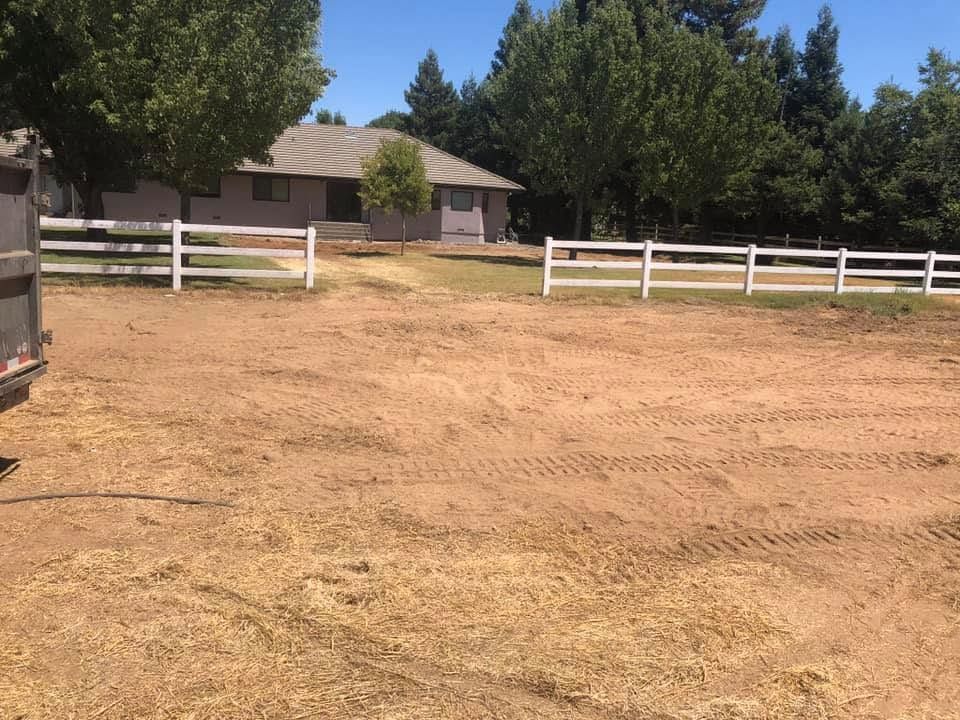 Dry dirt yard with white fences, a small house, and trees in the background.
