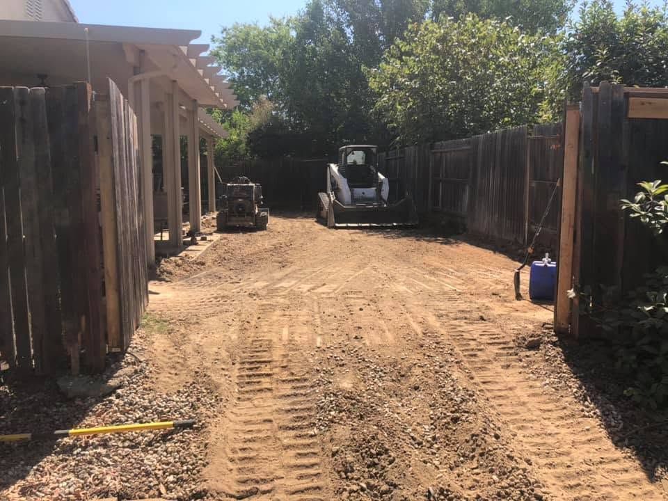 Dirt driveway between wooden fences, with a skid steer parked at the far end under trees.