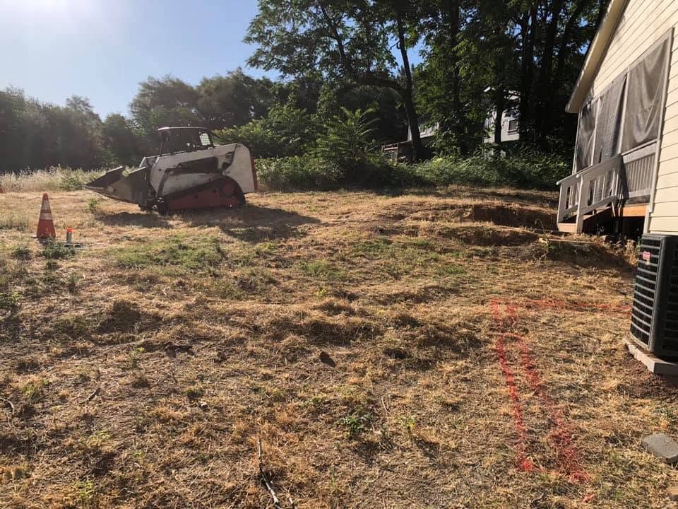 Construction site with excavator, orange cones, and a large truck on a cleared dirt lot near trees