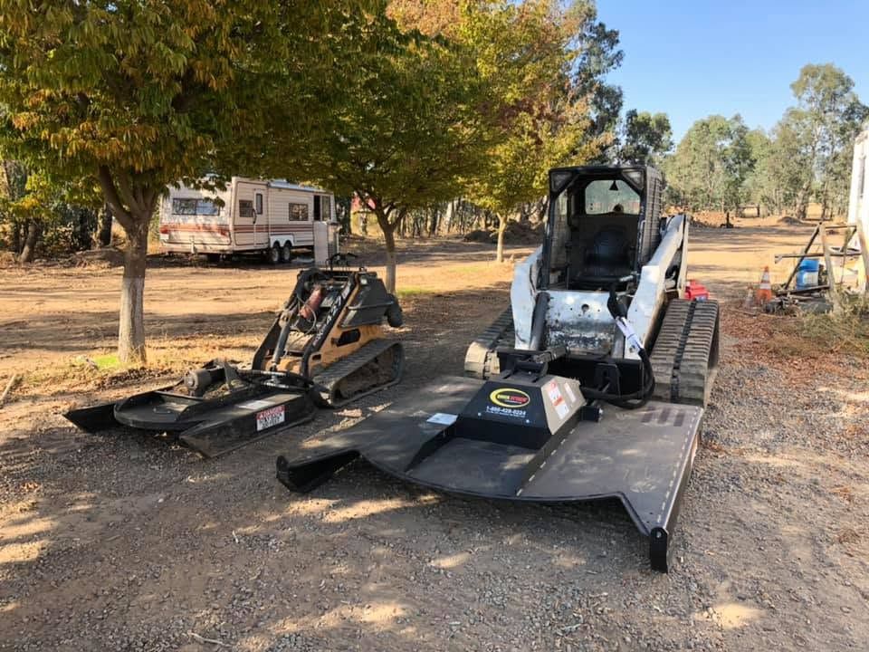Two compact track loaders with attachments parked in a dirt lot under trees, near a camper trailer.