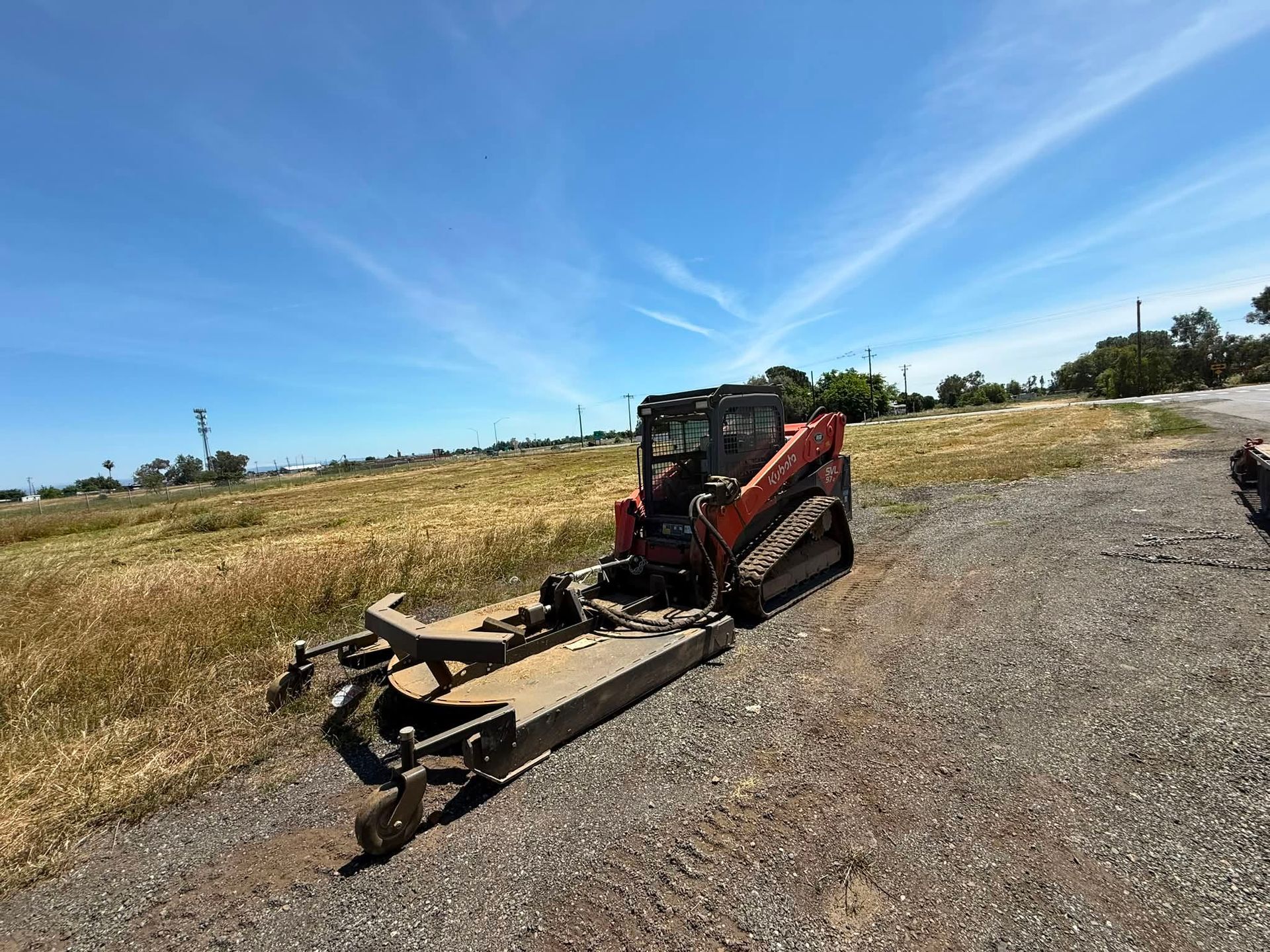 Red skid-steer loader with mower attachment on a gravel field under a blue sky