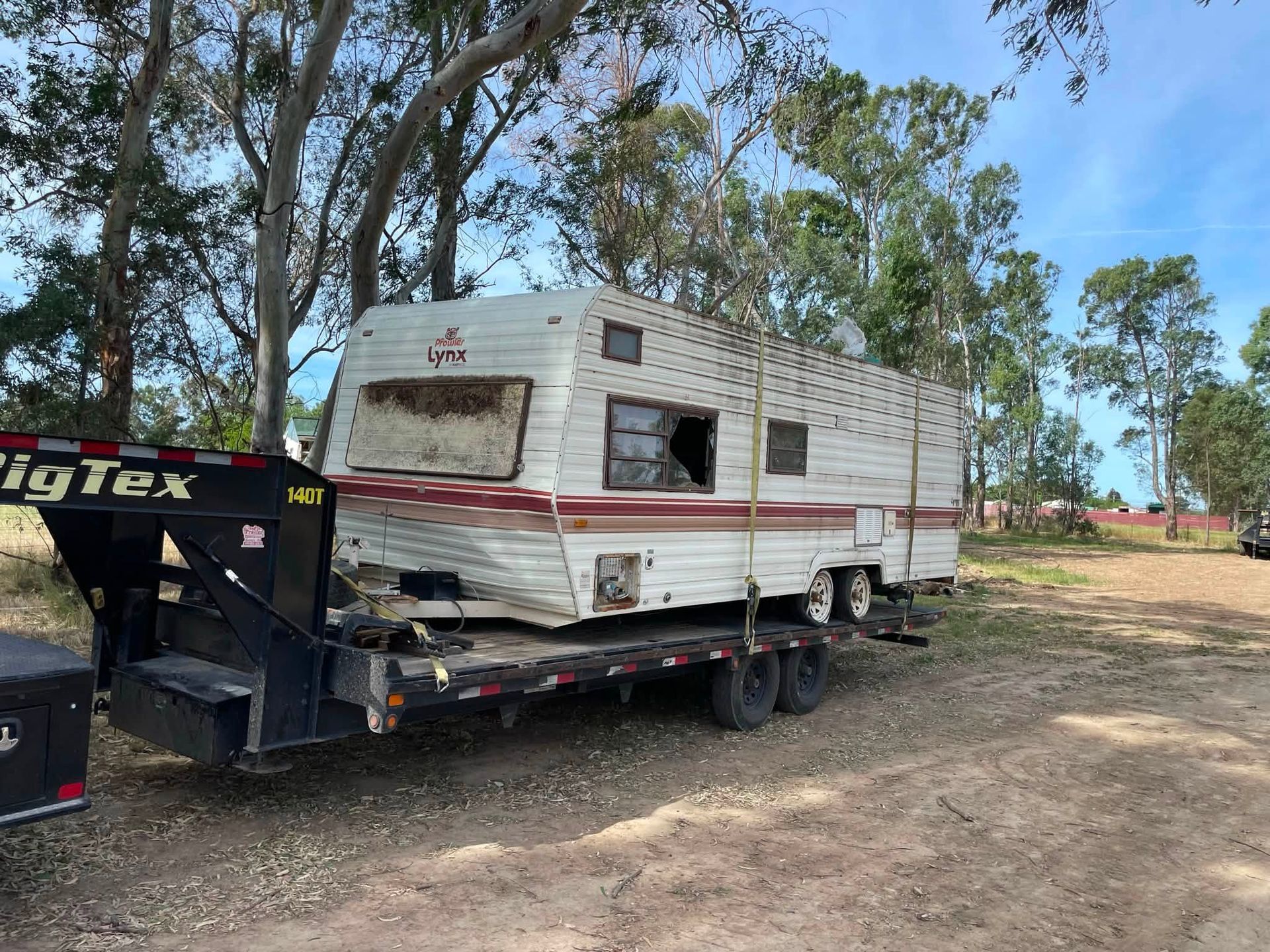 Damaged white camper trailer on a flatbed tow truck in a dirt clearing with trees behind it