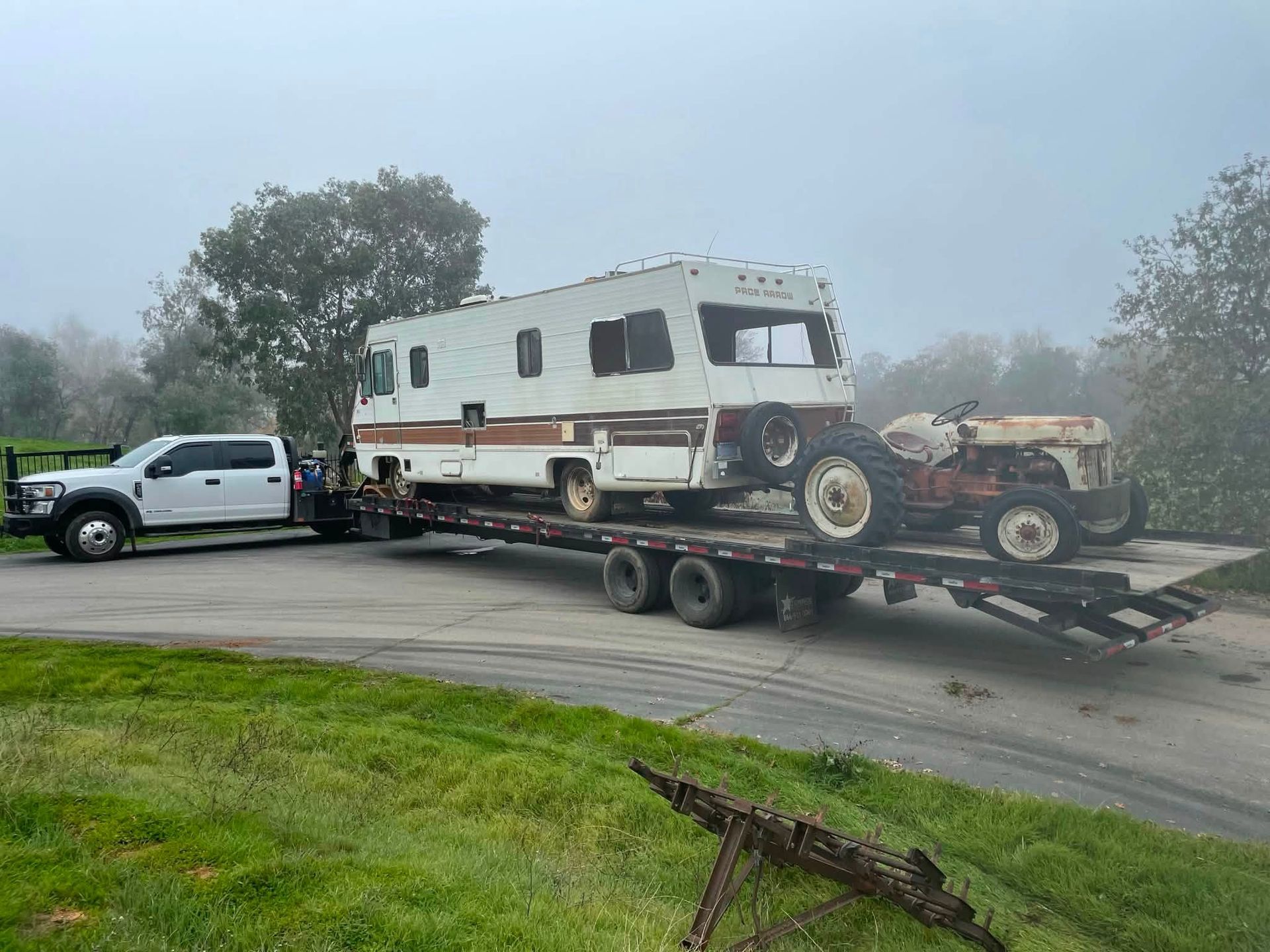White camper van on a trailer being towed by a pickup truck on a foggy road.
