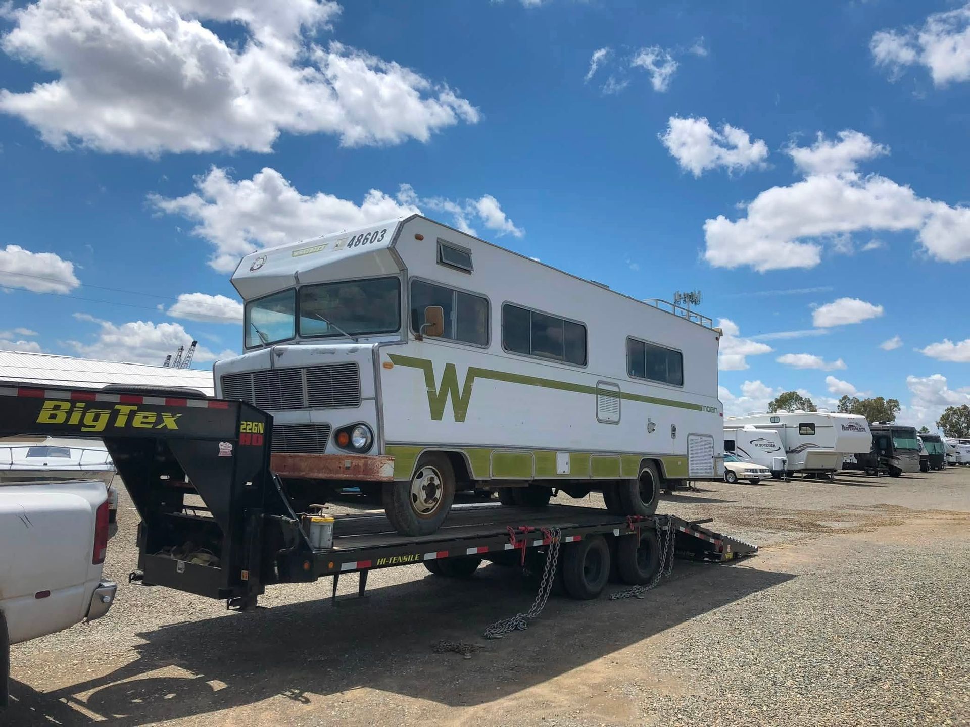 White RV on a tow truck in a gravel lot under a blue sky with clouds