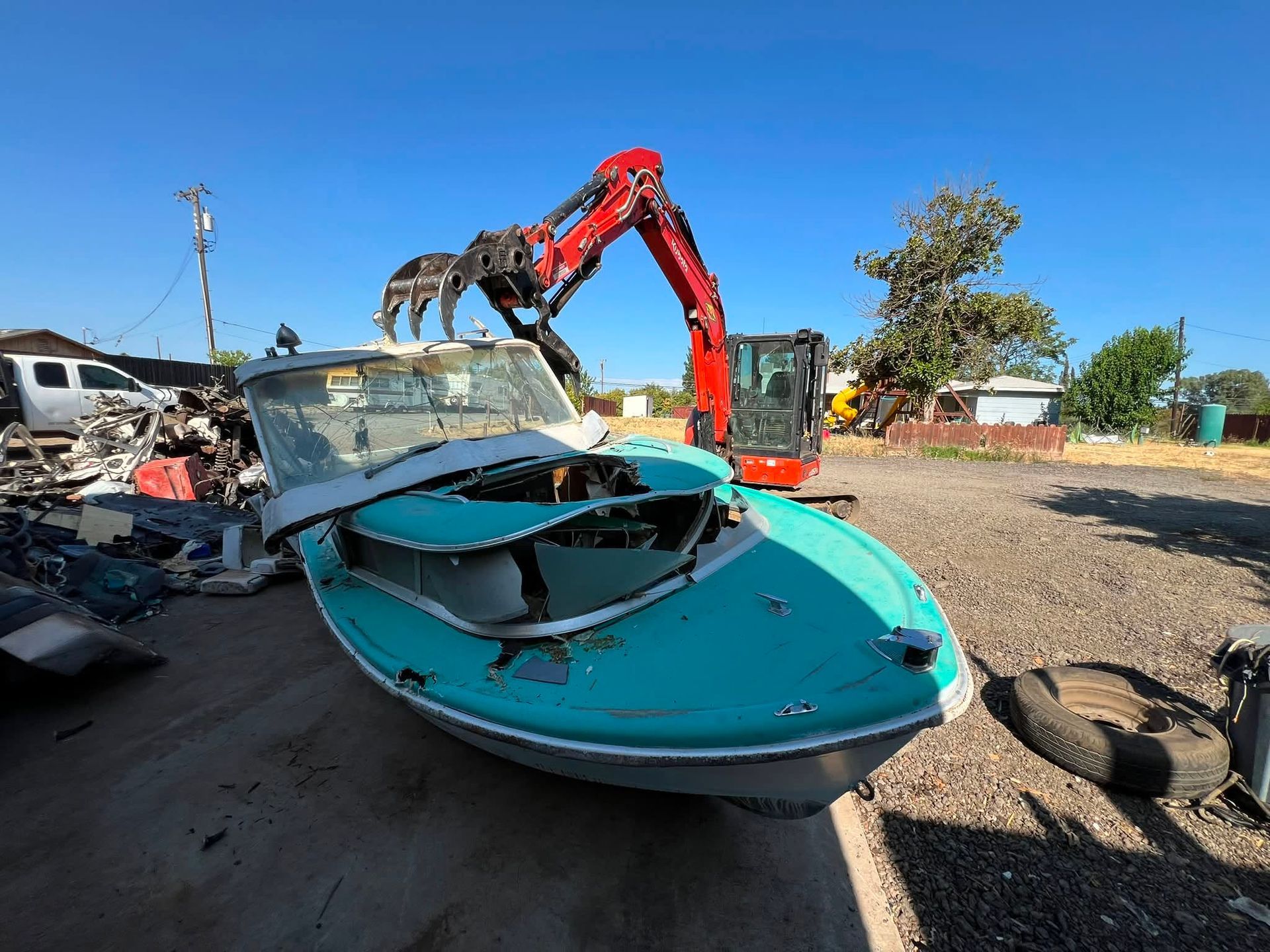Crushed turquoise boat at a scrapyard, with a red excavator lifting debris nearby.