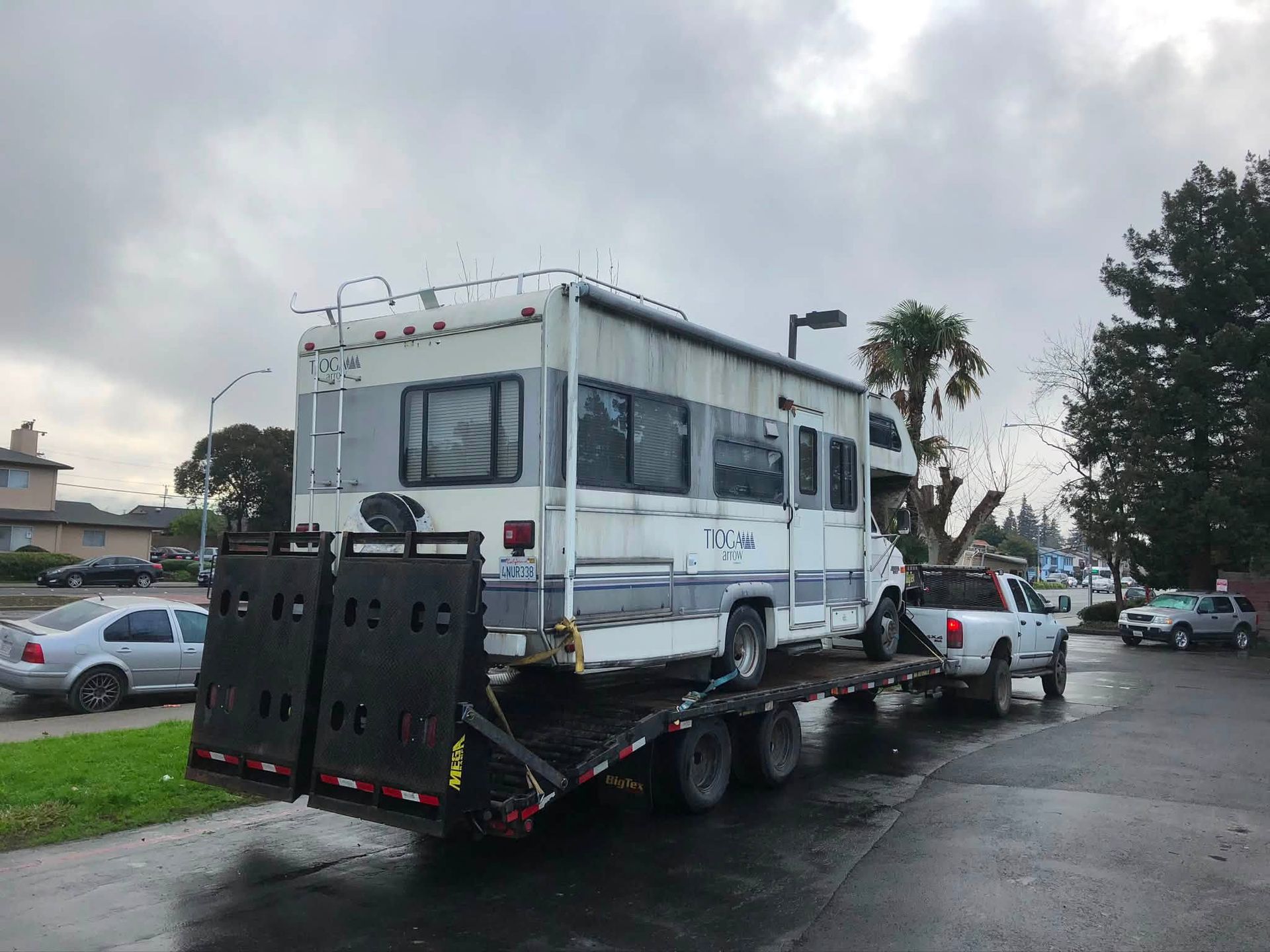 White RV loaded on a flatbed tow truck on a wet street under cloudy skies