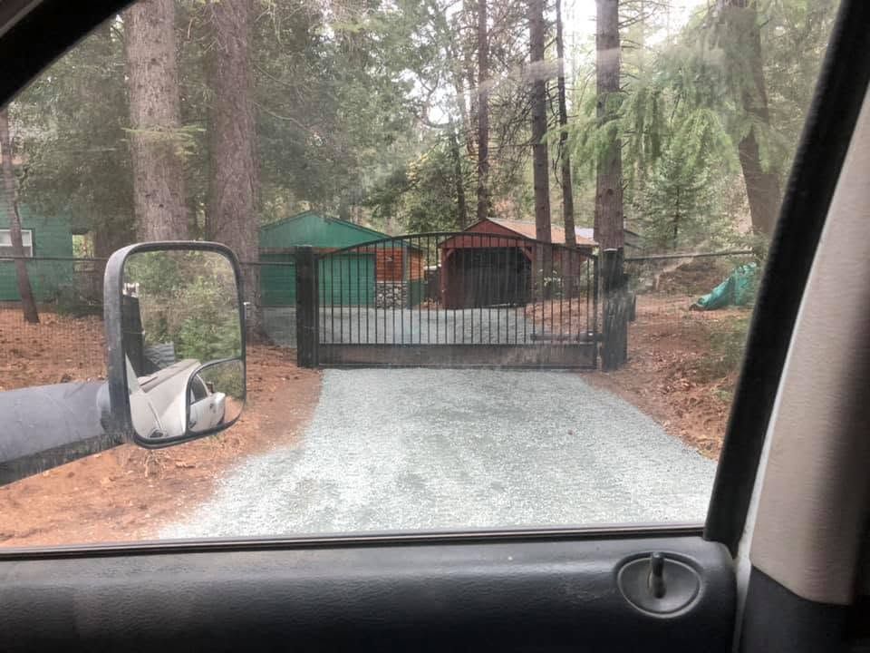Gravel driveway blocked by a black gate, seen from inside a vehicle in a wooded yard