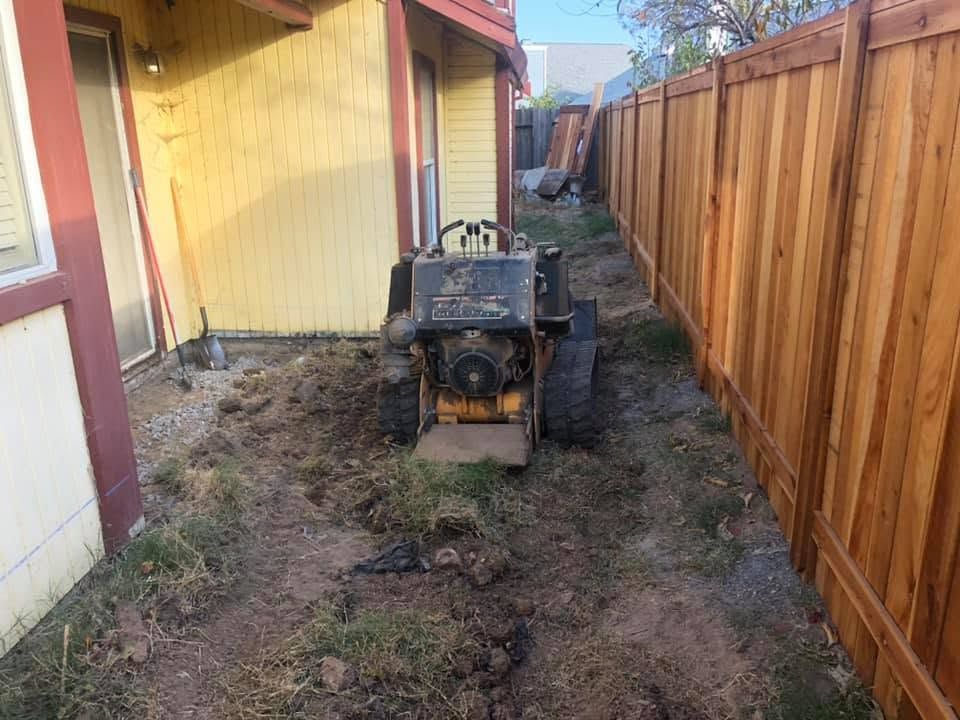 Small black tractor in a narrow muddy alley between a yellow house and a wooden fence.