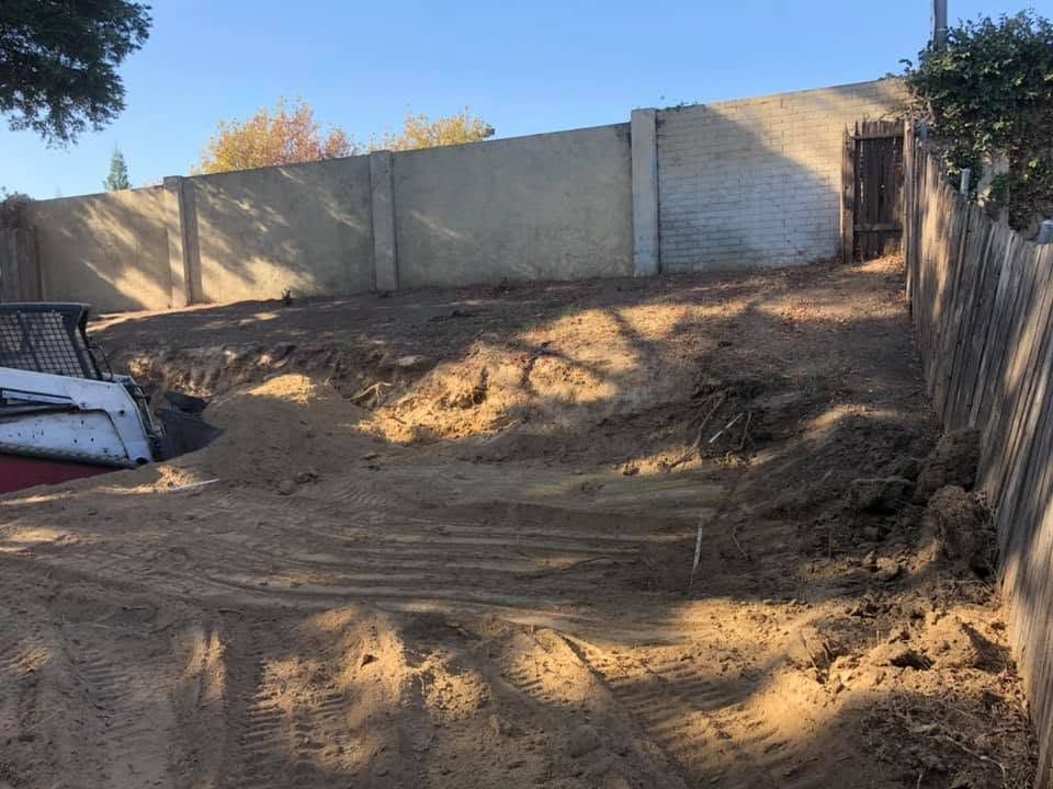 Empty dirt lot with sloped ground, wooden fence, and tall concrete wall in sunlight