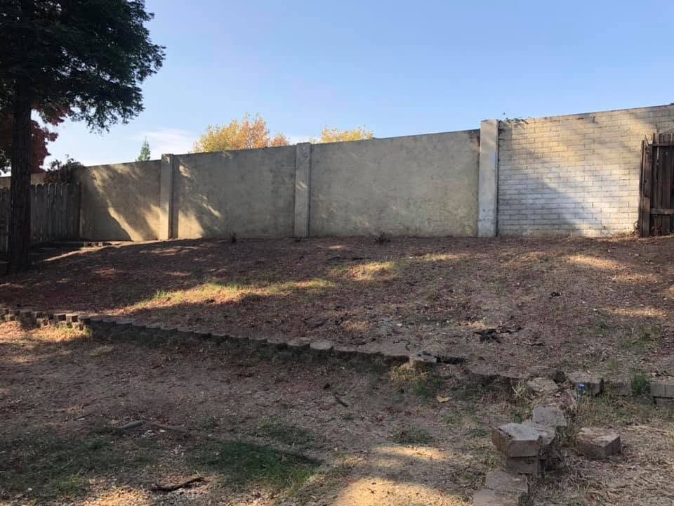 Empty dirt lot beside a tall concrete wall under a clear sky, with a tree casting shade on the left.