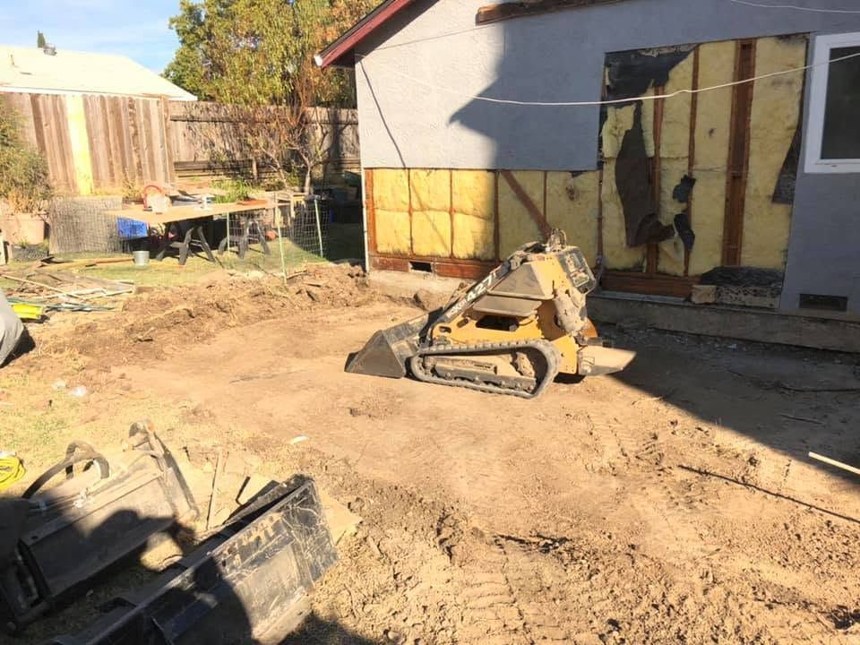 Backyard construction site with a small tracked loader and dirt piles beside a house wall