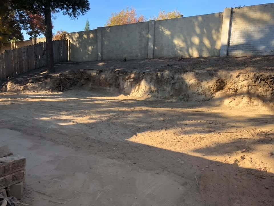 Dirt lot with a concrete wall, tree shadows, and exposed soil embankment in daylight