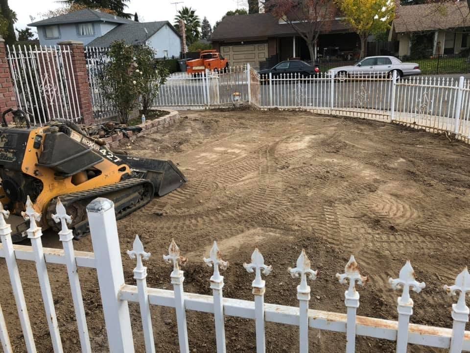 Backyard cleared for construction with a small excavator and dirt leveled inside a white picket fence