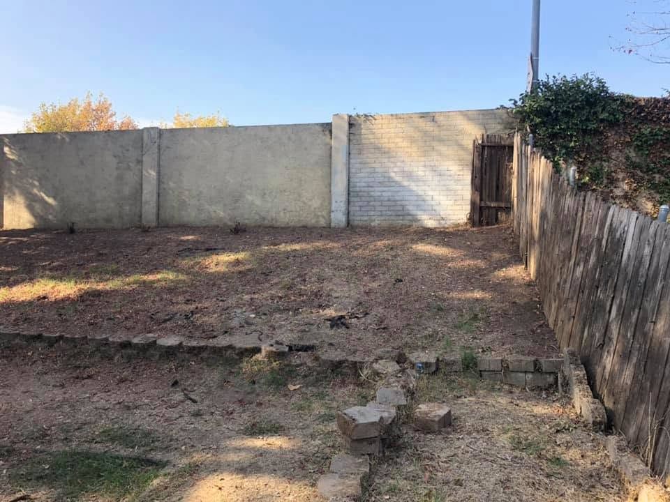 Empty dirt yard bordered by concrete walls and a wooden fence, with a small gate and dry grass.