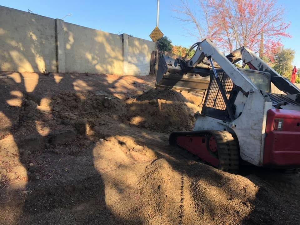 Skid-steer loader dumping dark soil into a pile beside a concrete wall outdoors.