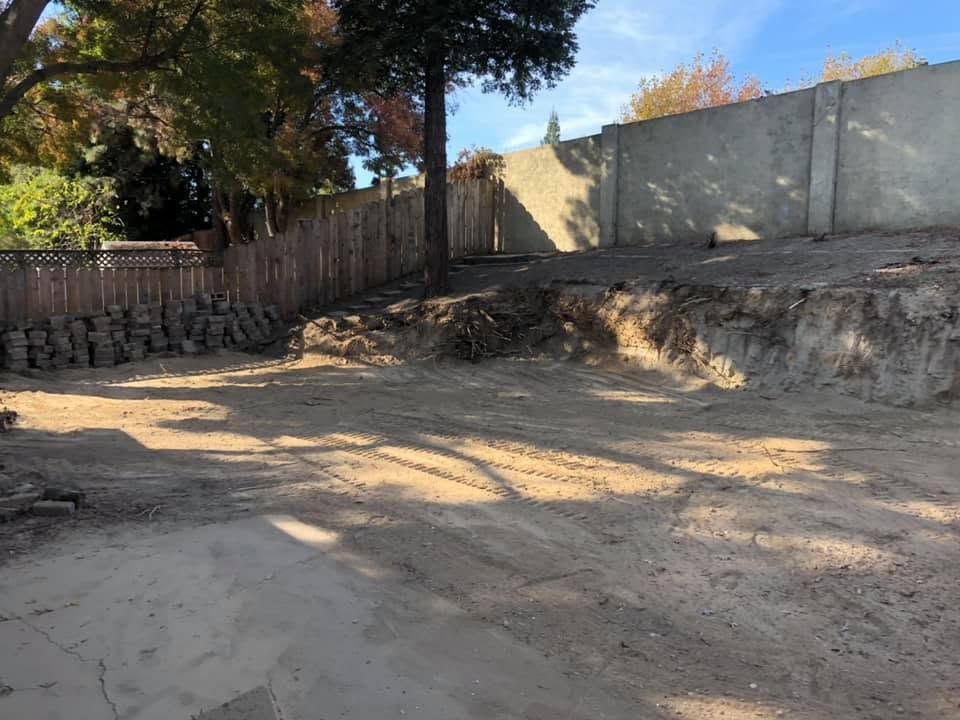 Empty dirt lot beside a wooden fence and concrete wall, with trees casting shadows.