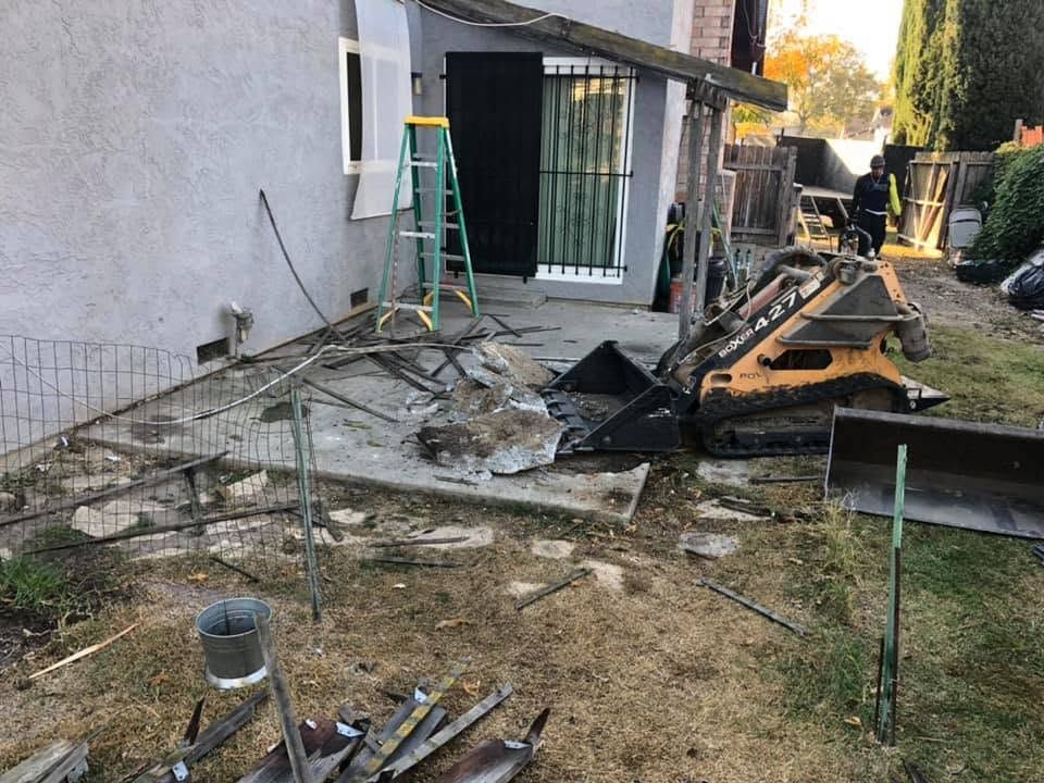 Backyard with construction debris, a skid steer, ladder, and damaged patio beside a house.