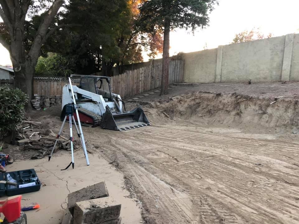 Backhoe on a dirt lot beside a tall retaining wall, with a tripod in the foreground.