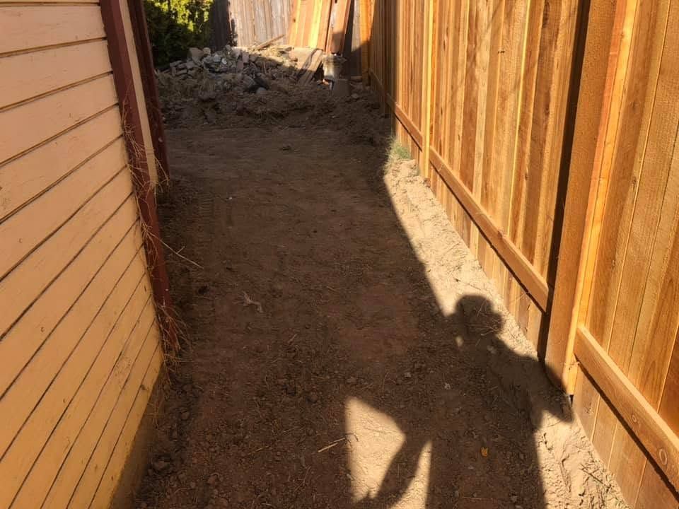 Narrow dirt alley between a house and wooden fence, with a person’s shadow on the ground.