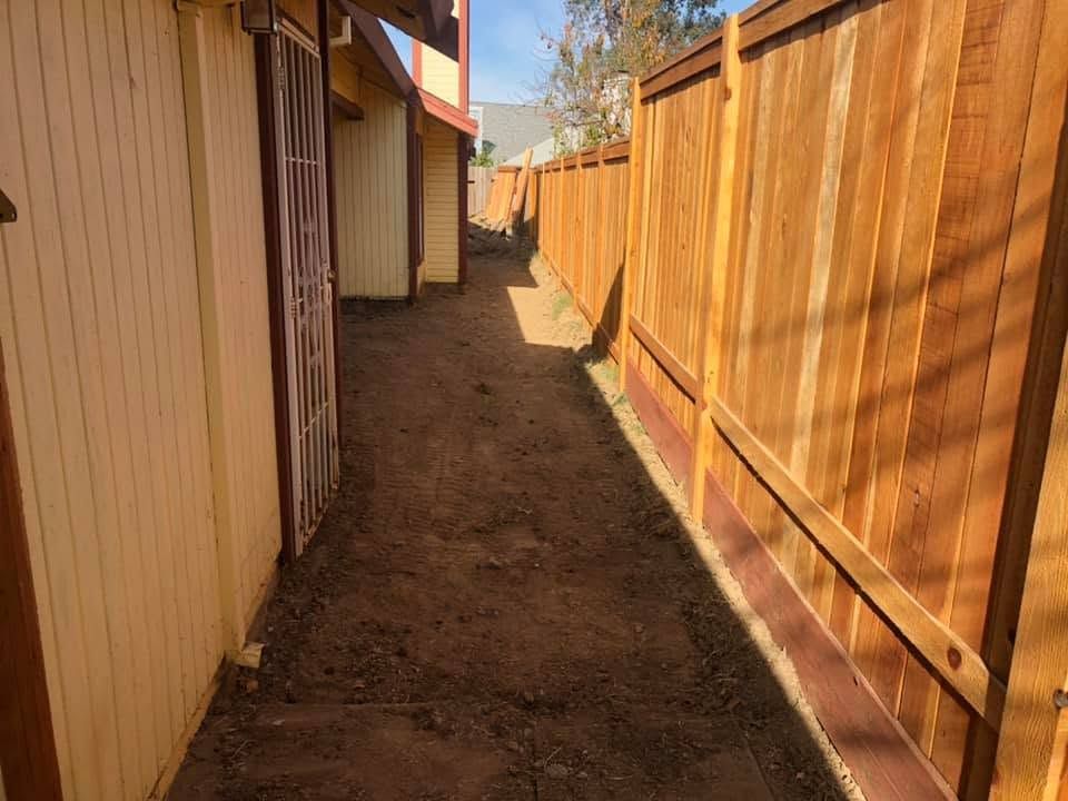 Narrow dirt walkway between beige siding and a tall wooden fence in sunlight