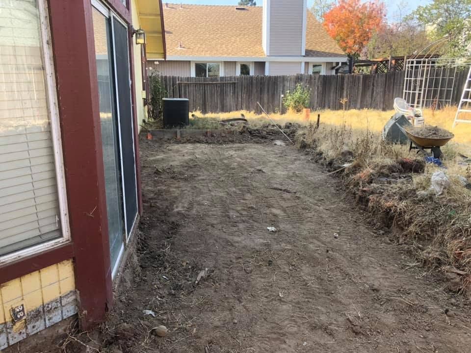 Narrow dirt side yard beside a house, with a fenced backyard and fall leaves in the distance.