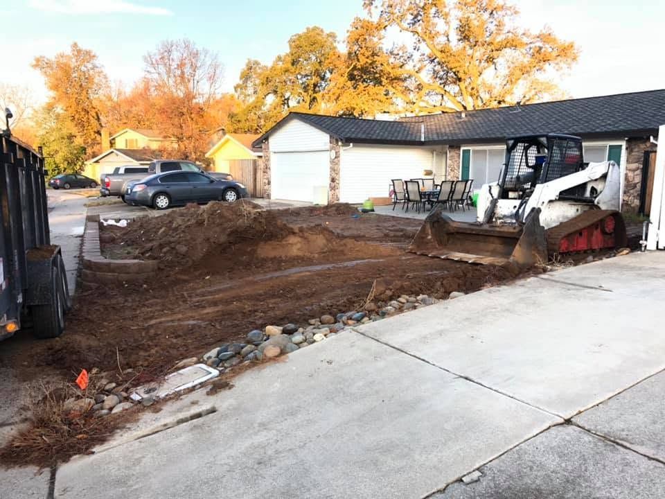 Backyard construction site with excavator, dirt trench, and a driveway beside a house and garage.