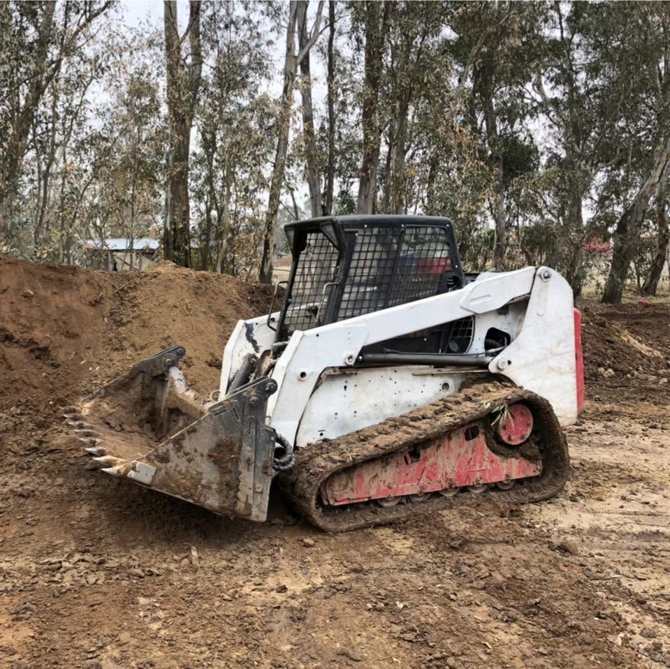 White skid-steer loader on muddy dirt road in a wooded area, with tracks covered in mud.
