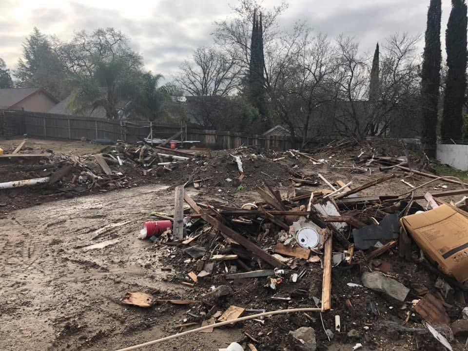 Debris and splintered wood scattered across a destroyed yard after a fire or storm, with damaged trees and homes in background