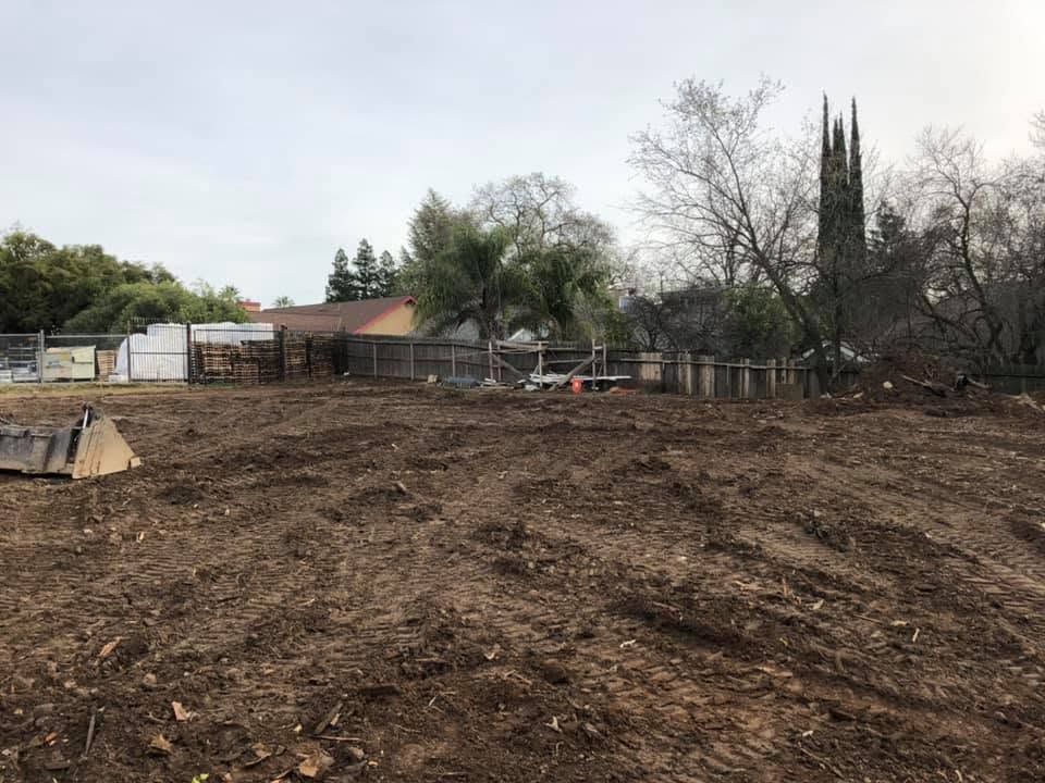 Cleared dirt lot with a fence and trees beside a small house under an overcast sky