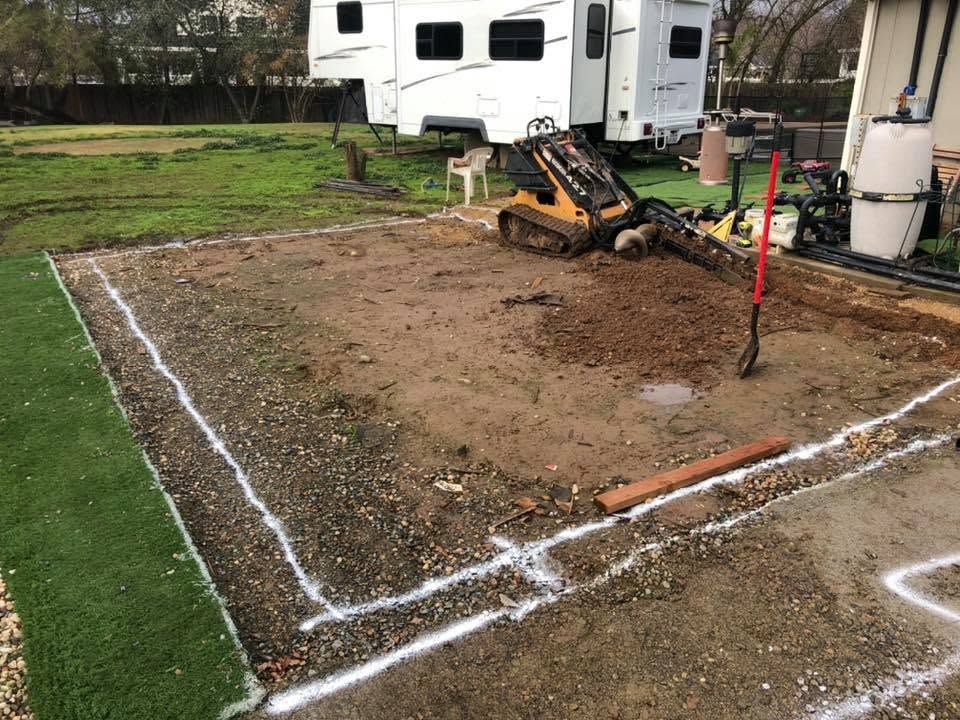 Marked dirt lot beside a trailer, with a small backhoe digging and white chalk outlines on the ground