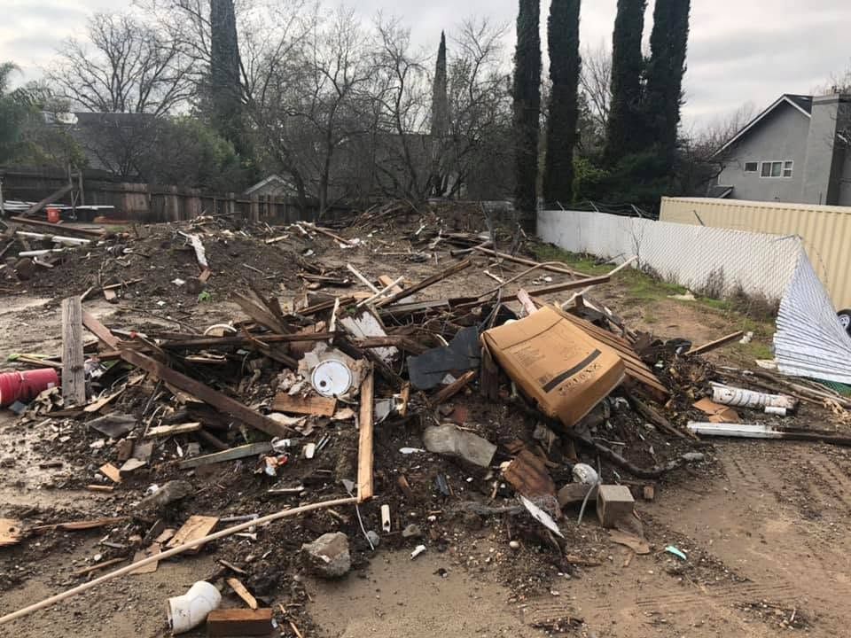 Debris and wreckage scattered across a muddy lot beside a white fence and houses after destruction