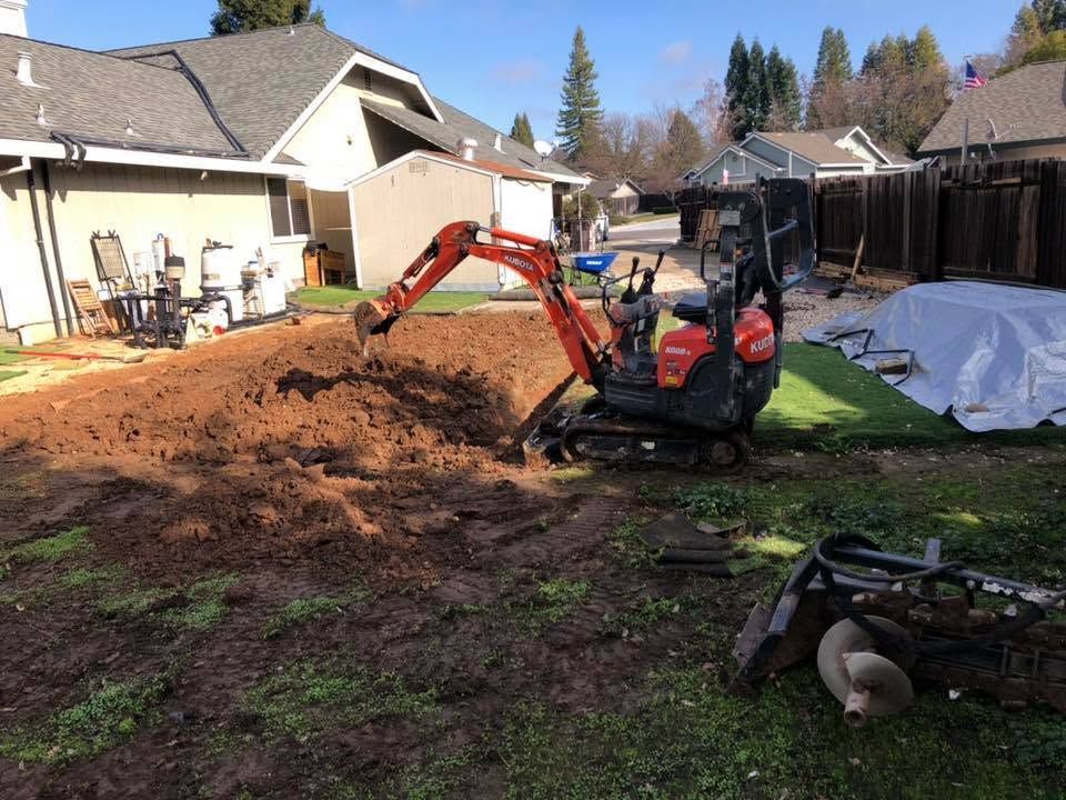 Mini excavator digging a large dirt pile in a suburban backyard with houses nearby